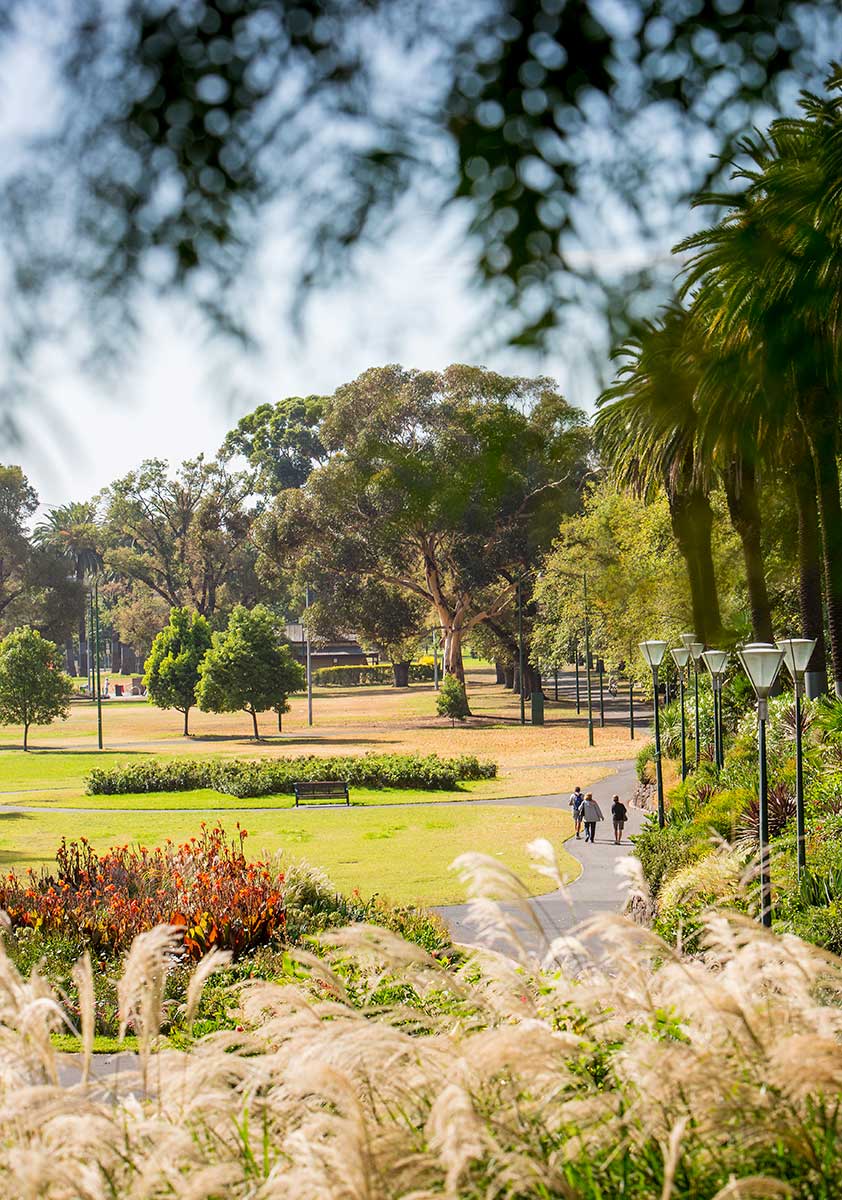 People walking on a footpath in Alexandra Gardens with adjacent grassland,  garden beds with flowers, shrubs and grasses, and mature trees.