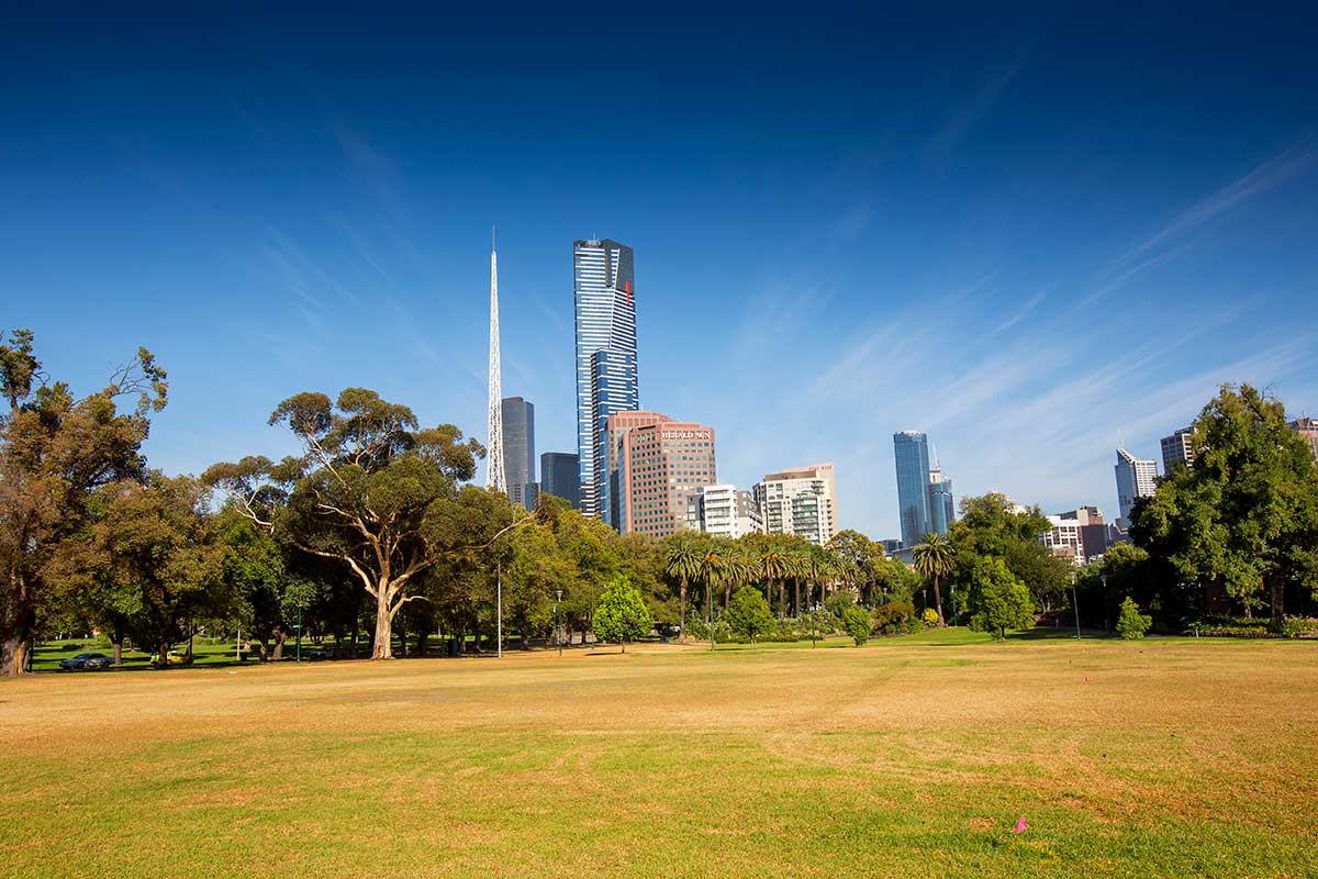 View of open grassland in Alexandra Gardens with a range of trees in the middle distance, and CBD buildings (including Eureka Tower and Arts Centre spire) in the background.