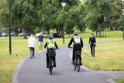 Two animal management officers riding bicycles on a path through a park cycle past two people walking dogs and a person walking alone