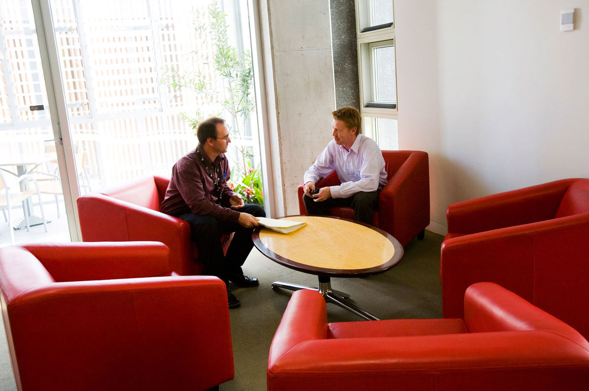 Two people using the breakout space. The space has large padded red armchairs around a small table, and is next to a large window