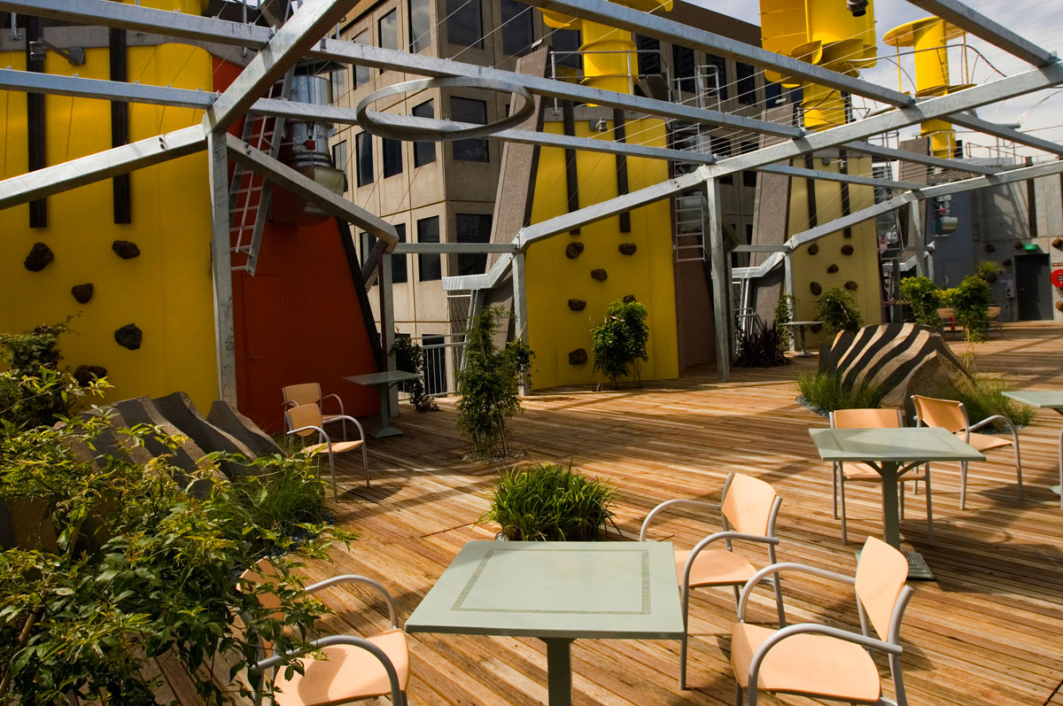 View of the building's rooftop showing large yellow turbines, tables and chairs, open areas with plants and a metal pergola-like structure