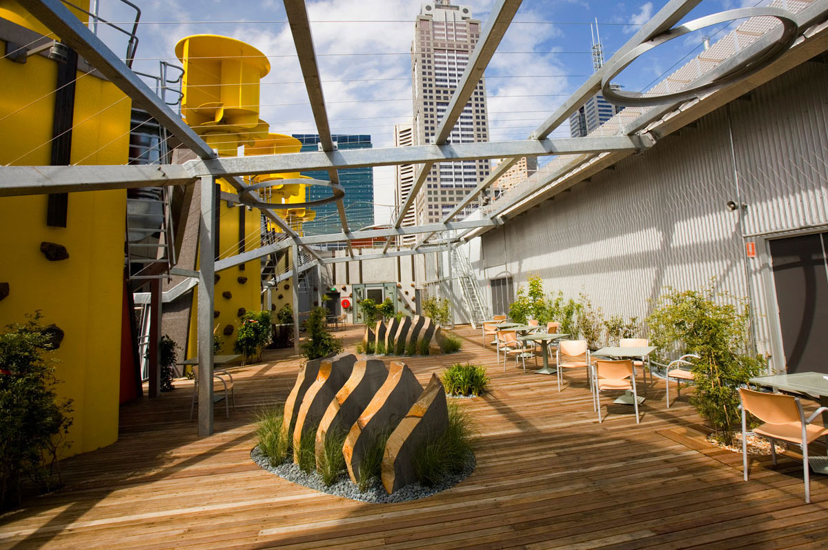 View of the building's rooftop showing the large yellow turbines, tables and chairs, plants and sculptures underneath the pergola structure