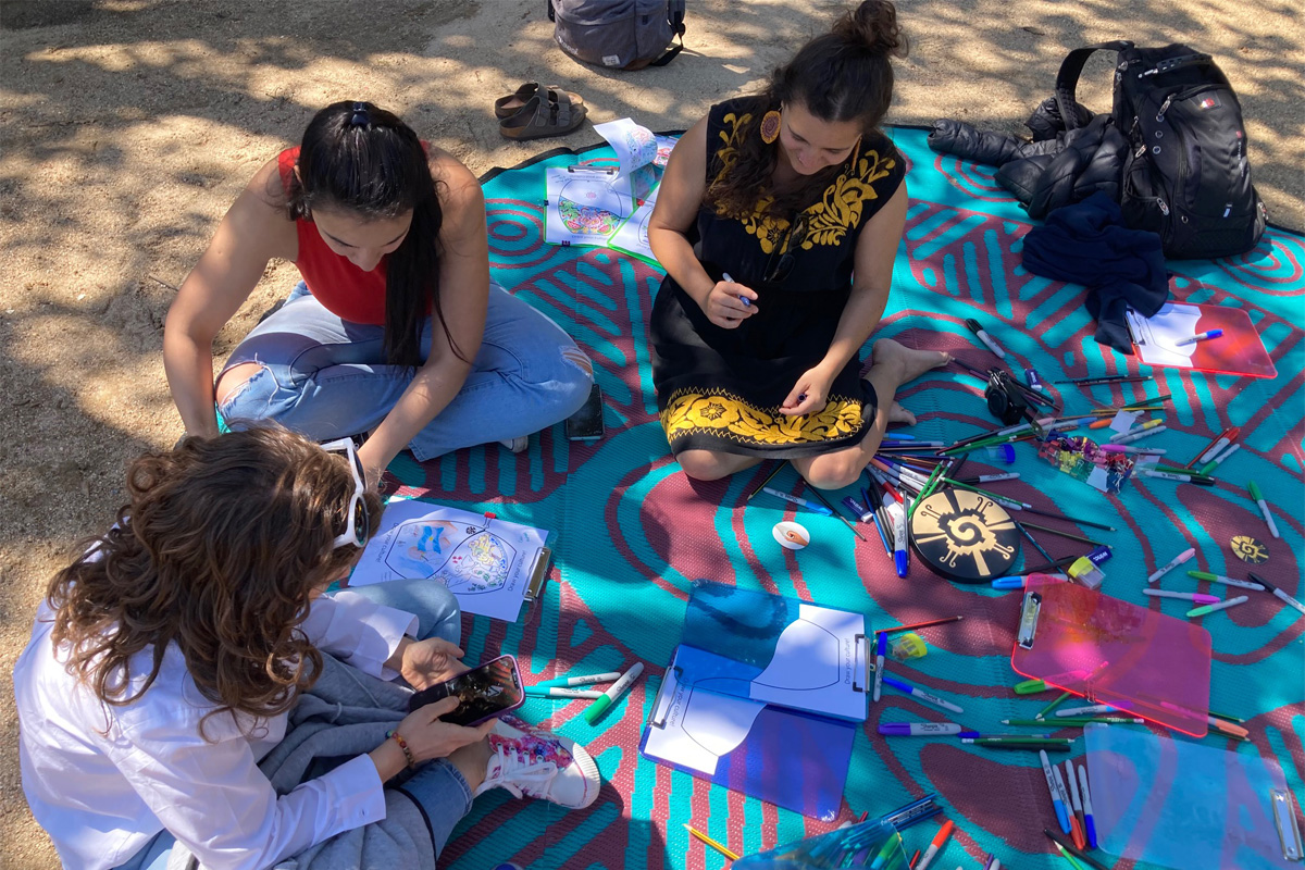 Three people sitting on a picnic rug with craft materials spread out around them.