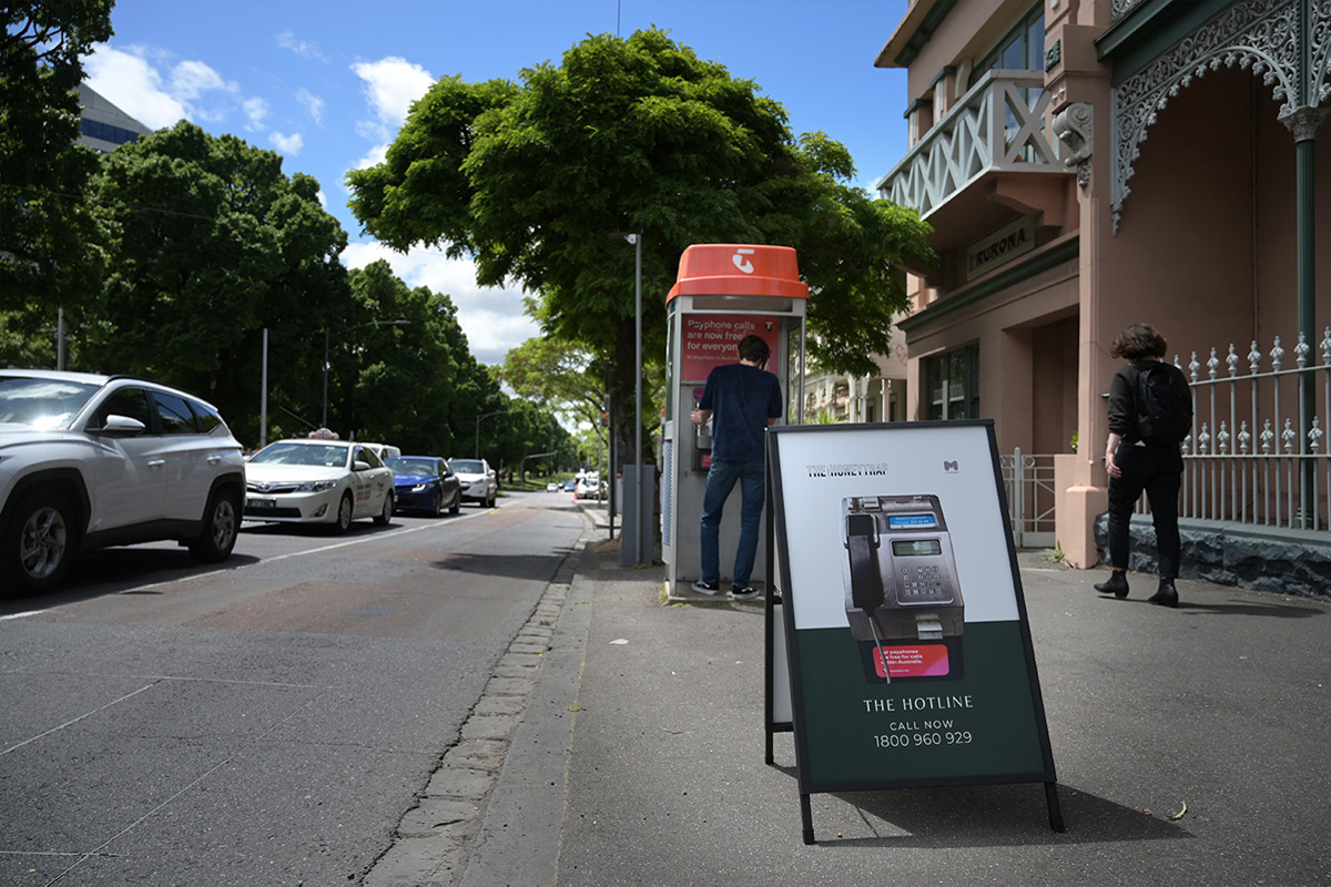 A sign on a street with an image of a public telephone on it that reads “The hotline call now 1800 960 929”.